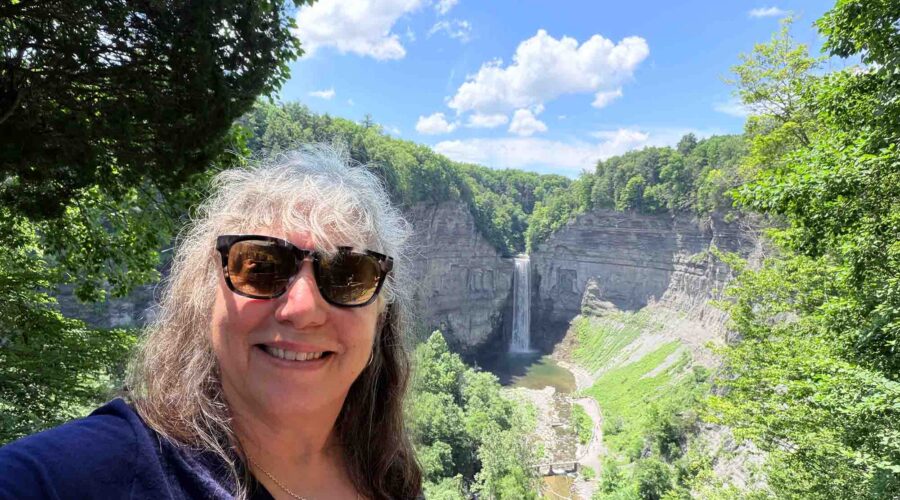 Woman taking a selfie outdoors with a waterfall and lush green trees in the background on a sunny day